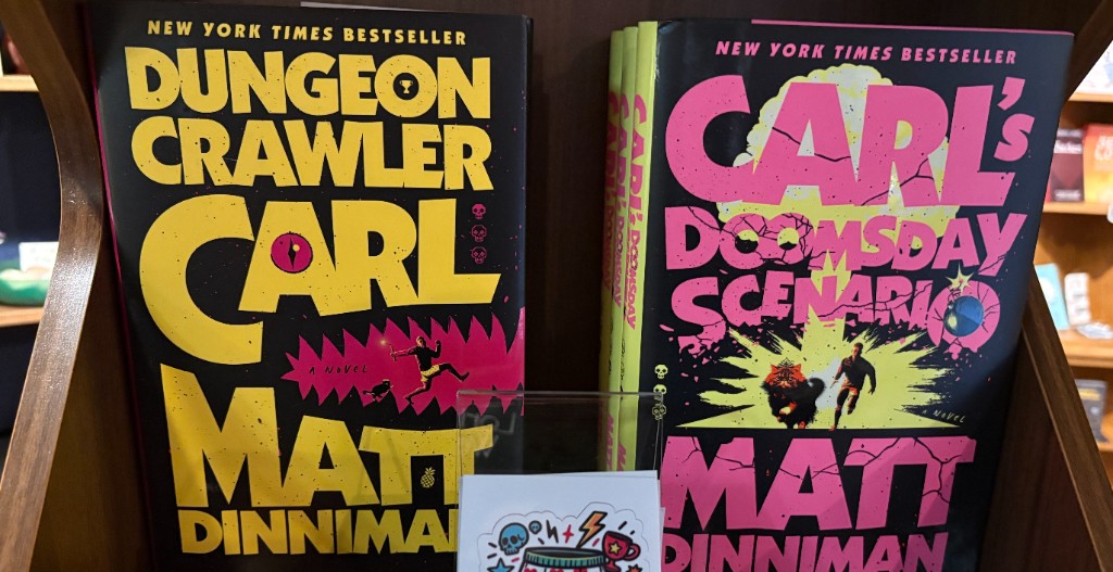 Dungeon Crawler Carl books and a “You Will Not Break Me” sticker on a shelf at Dragonfly Bookshop.